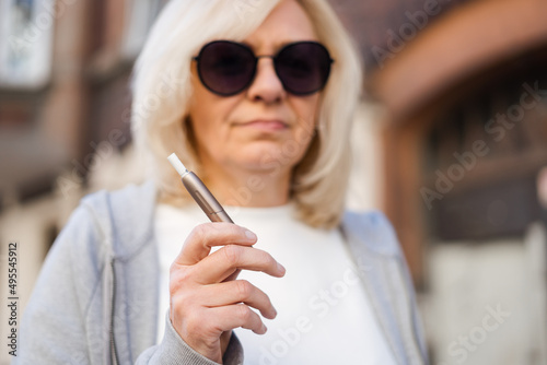 Middle aged blonde European woman smoking heated tabacco e-cigarette outdoors on the street. Alternative smoking way photo