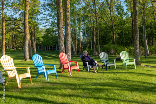 A woman sitting on an Adirondack chair with trees and woods in the background, on a sunny day with shadows.
