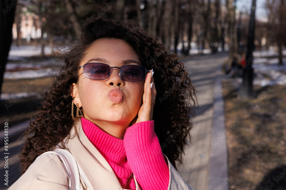 Happy curly brunette girl in sun glasses smiling outdoors. Young woman happy walking in street. Pink sweater, beige coat, beige hat. Sun in city. Fashionable asian girl with frizzly hair