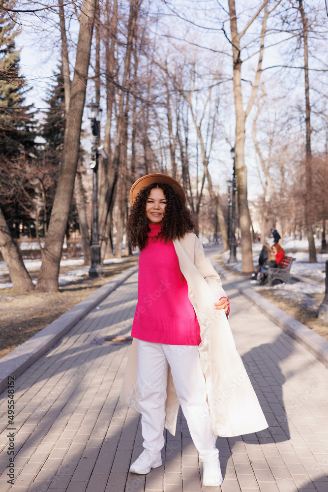 Happy curly brunette girl in hat smiling outdoors. Young woman happy walking in street. Pink sweater, beige coat, beige hat. Sun in city. Fashionable asian girl with frizzly hair Happy curly brunette girl in hat smiling outdoors. Young woman happy walking in street. Pink sweater, beige coat, beige hat. Sun in city. Fashionable asian girl with frizzly hair