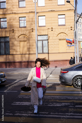 Happy curly brunette girl smiling outdoors. Young woman happy walking in street. Pink sweater, beige coat, beige hat. Sun in city. Fashionable asian girl with frizzly hair © Tatyana