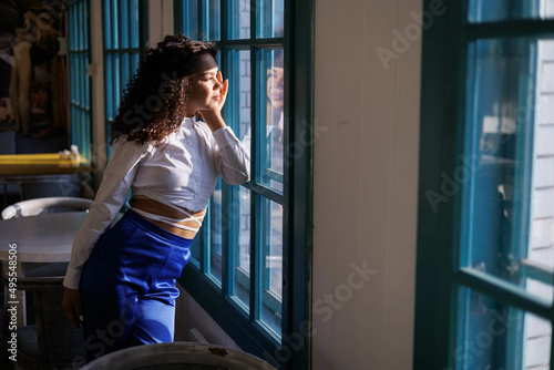 Happy curly brunette girl smiling in cafe. Young woman enyoing sun in restaurant at window. white blouse. Sun in city. Fashionable asian girl with frizzly hair. Sun light in window