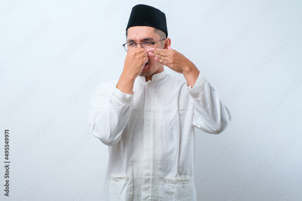 Asian muslim man wearing glasses over white background smelling something stinky and disgusting Asian muslim man wearing glasses over white background smelling something stinky and disgusting