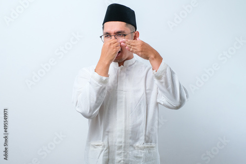 Asian muslim man wearing glasses over white background smelling something stinky and disgusting