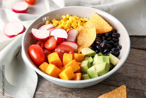 Bowl of Mexican vegetable salad with black beans, radish and nachos on wooden background, closeup