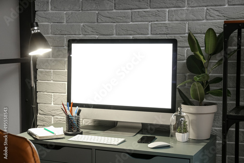 Workplace with modern computer, mobile phone, stationery supplies and glowing lamp near grey brick wall