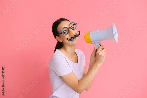 Young woman with funny disguise shouting into megaphone on pink background. April Fools Day celebration