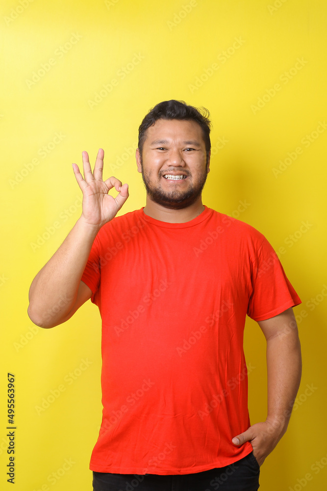Excited Asian man in basic clothing smiling and showing ok sign at camera isolated over yellow background Excited Asian man in basic clothing smiling and showing ok sign at camera isolated over yellow background
