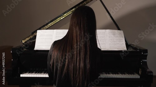 Brunette woman in black clothes plays the piano alone in the hall. Sheets of paper with notes are on the music stand