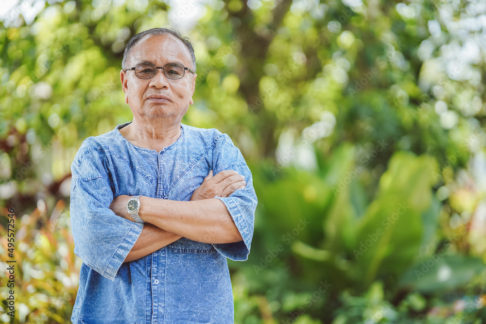 Fototapeta premium An elderly Thai man standing with his arms crossed looking at the camera with a blurred natural background. Copy space.
