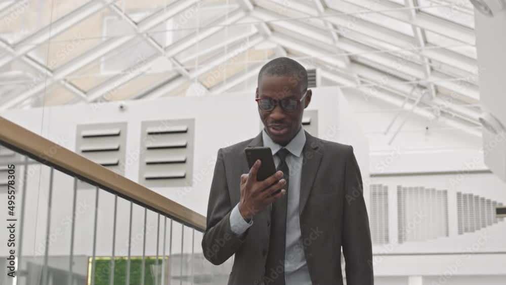 Tracking medium slowmo of young confident African American in formal wear and eyeglasses talking on cellphone while walking along modern business centre Tracking medium slowmo of young confident African American in formal wear and eyeglasses talking on cellphone while walking along modern business centre