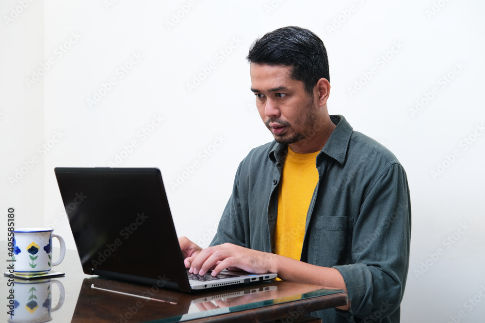 Adult Asian man showing serious expression when typing on his laptop Adult Asian man showing serious expression when typing on his laptop