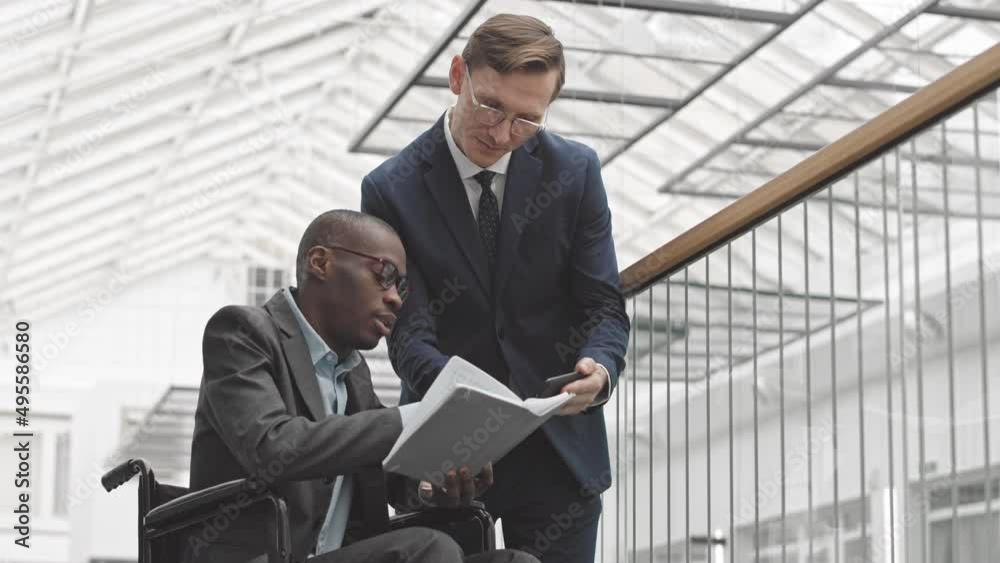 Medium slowmo of successful Black businessman in wheelchair holding notebook with project strategy discussing it with Caucasian business partner standing nearby. Men shaking hands in agreement Medium slowmo of successful Black businessman in wheelchair holding notebook with project strategy discussing it with Caucasian business partner standing nearby. Men shaking hands in agreement