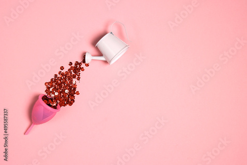 Red sparkles are poured into the menstrual cup from a small watering can. Pink background. photo