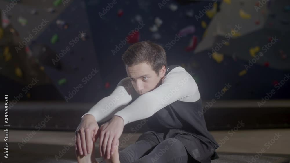 Young male athlete doing stretching warm-up exercises indoors. Sportsman touching toes with his hands before training in bouldering gym. Artificial rock wall in background. Sport, bouldering concept Young male athlete doing stretching warm-up exercises indoors. Sportsman touching toes with his hands before training in bouldering gym. Artificial rock wall in background. Sport, bouldering concept