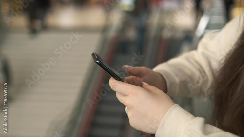 Hands of a young woman using a smartphone in a shopping mall. A young woman is holding a phone in a shopping mall in front of an escalator with people. Close up photo