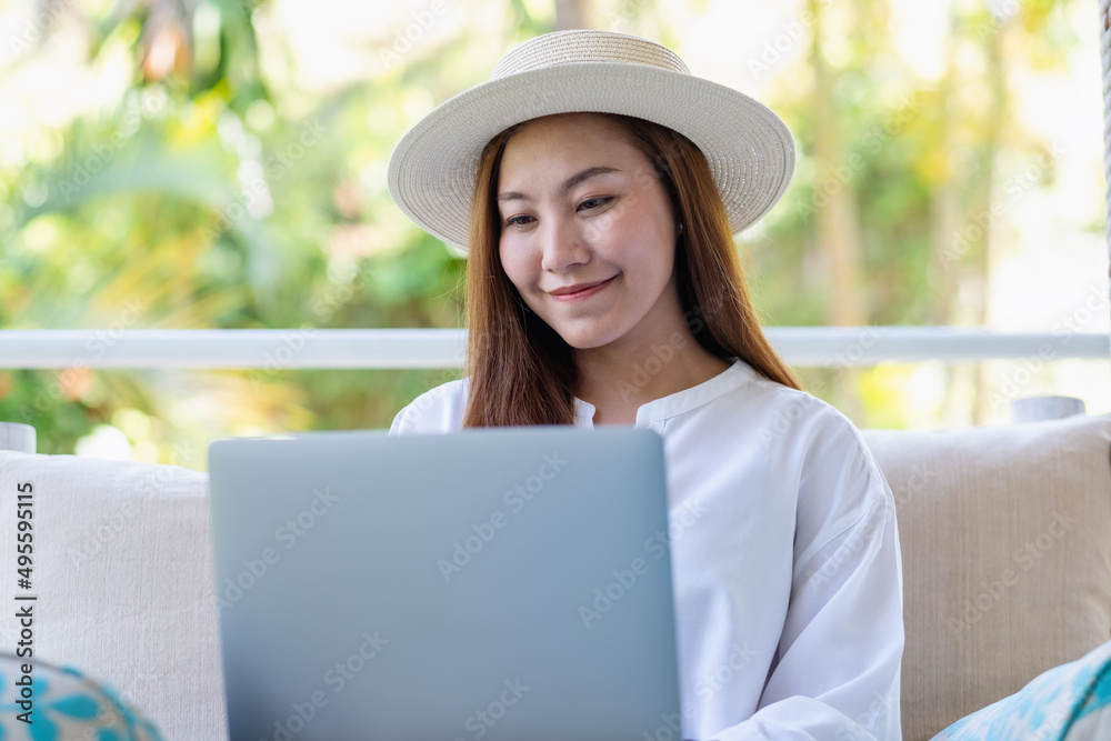 Portrait image of a beautiful young asian woman using laptop computer for remote working or studying online while sitting on swing sofa