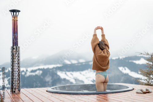 Woman going to rest in hot tub on terrace with great view on snowy mountains. Concept of rest and recovery in hot vat on nature. Idea of escape and recreation on mountains