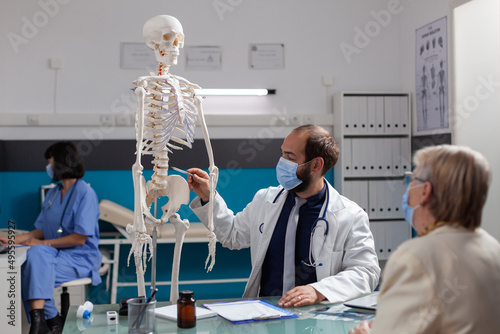 Specialist explaining human skeleton to retired woman at checkup visit, talking about bones injury at osteopathy appointment during covid 19 pandemic. Doctor showing joint model to patient.