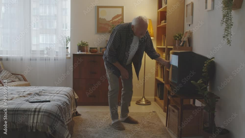 Irritated retired Caucasian man sitting on bed at home in evening, then standing up, checking old broken CRT TV Irritated retired Caucasian man sitting on bed at home in evening, then standing up, checking old broken CRT TV