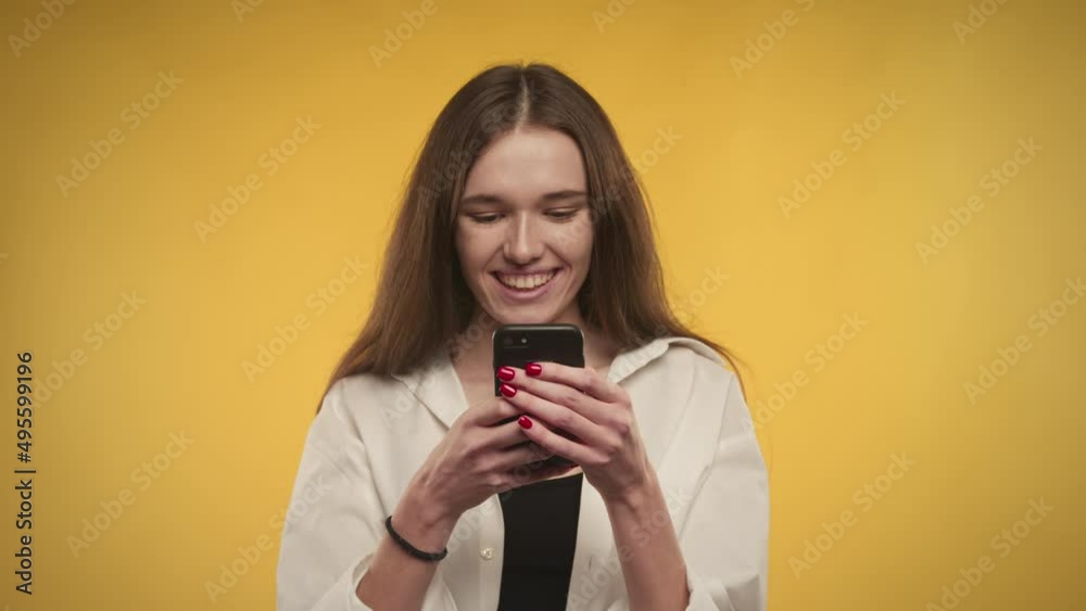 Young adult caucasian woman is happy texting on her phone on a bright yellow background