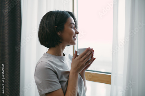 Smiling beautiful young woman looking out window with cup of hot drink relaxing in morning at home. Domestic life