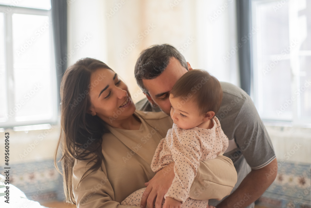 Portrait of happy mid adult parents and baby son at home. Mother holding child in arms and father kissing him. Parenthood concept Portrait of happy mid adult parents and baby son at home. Mother holding child in arms and father kissing him. Parenthood concept