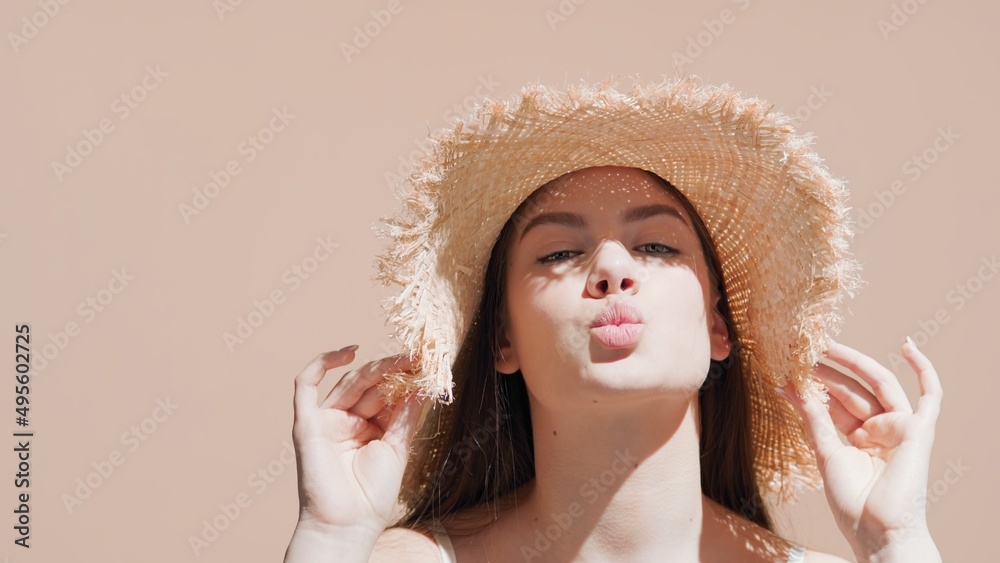 Young attractive European woman with long brown hair in a straw hat holding the brim of it blows a kiss for the camera and smiles touching her shoulder against beige background | Sunblock commercial Young attractive European woman with long brown hair in a straw hat holding the brim of it blows a kiss for the camera and smiles touching her shoulder against beige background | Sunblock commercial