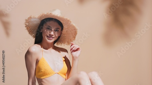 Young brunette asian woman in straw hat and yellow crochet bikini enjoys the sun, sunbathing and smiles for the camera | Sunscreen protection and skin care commercial concept