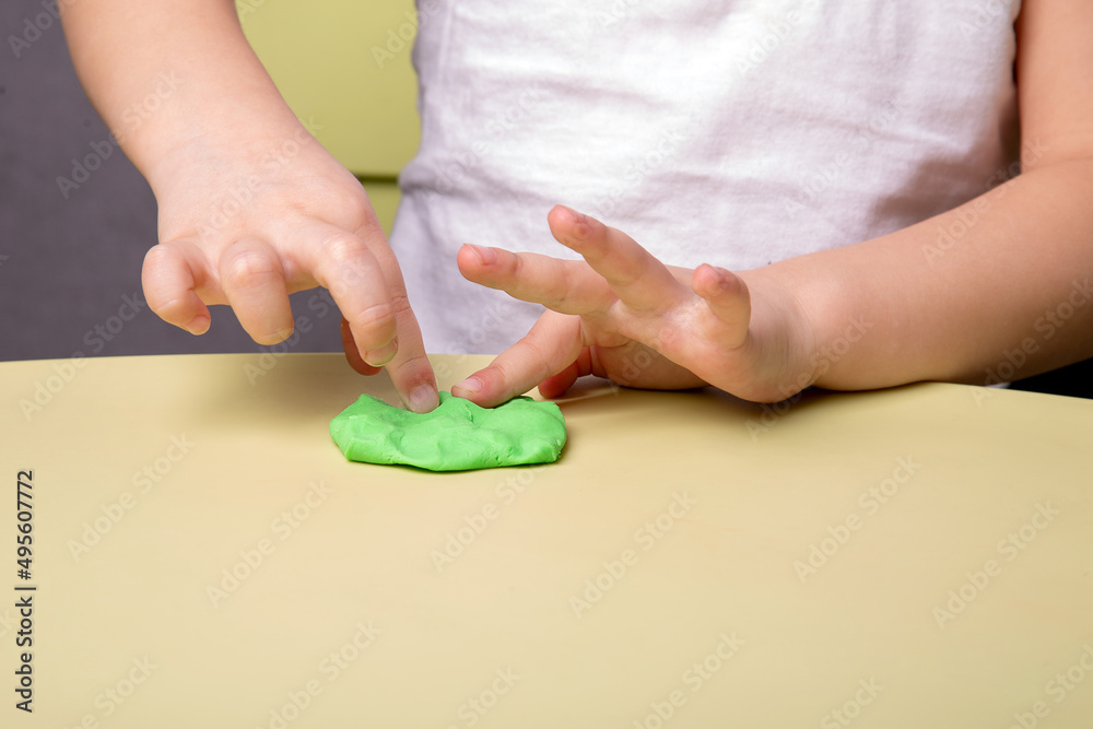 A child crumples a piece of green plasticine on the table, close-up of hands A child crumples a piece of green plasticine on the table, close-up of hands