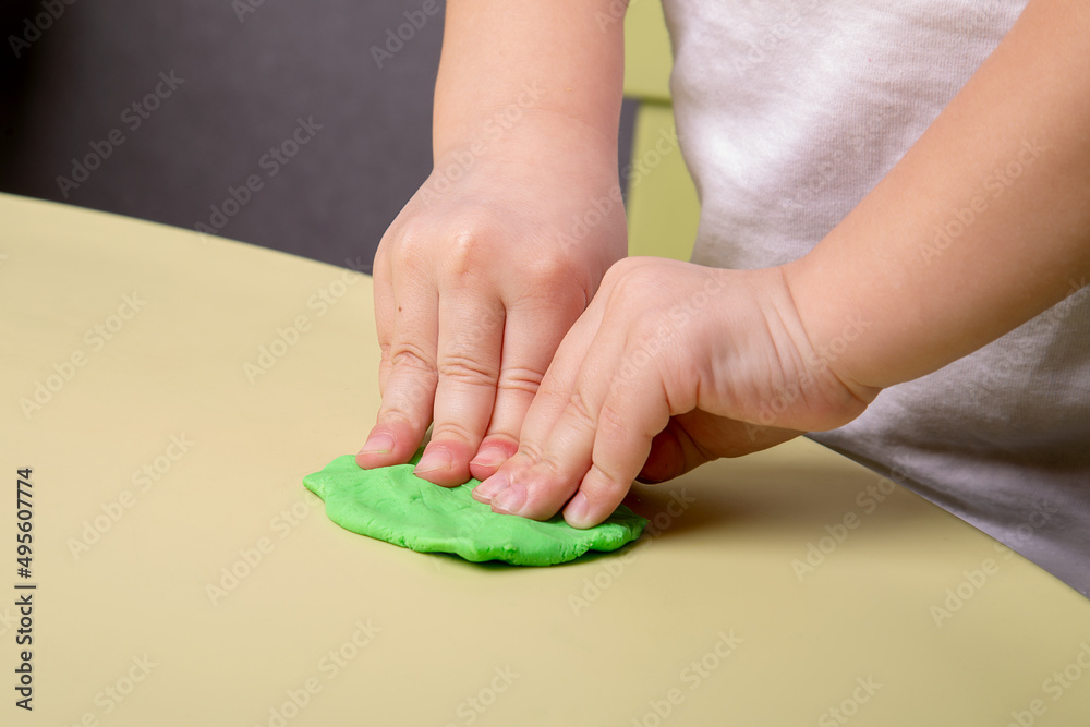 A child crumples a piece of green plasticine on the table, close-up of hands A child crumples a piece of green plasticine on the table, close-up of hands