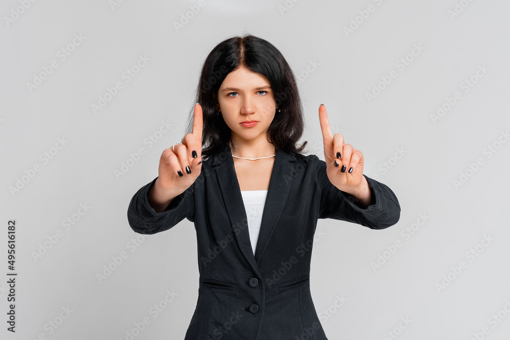 Please, do not, stop. Young serious dark-haired girl in black blazer, who is looking in the camera and pointing fingers up, standing over grey background Please, do not, stop. Young serious dark-haired girl in black blazer, who is looking in the camera and pointing fingers up, standing over grey background