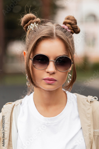 Fashion portrait of beauty young woman model with round retro sunglasses and hairstyle in stylish sports clothes with windbreaker and white t-shirt on the street