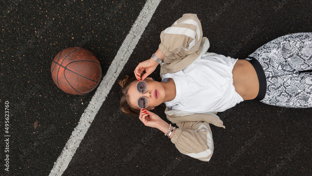 Fototapeta premium Sporty beautiful sexual woman hipster in fashionable sports clothes with a ball lying on the pavement and wearing fashion sunglasses, top view. Girl rest on the street