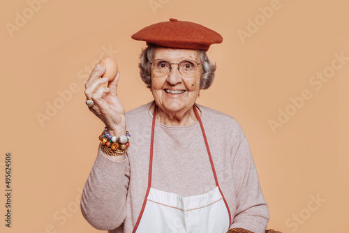 Happy smiling elderly grandmother cook holding an egg, looking at camera. Grey-haired old cook wearing apron and hat at studio over beige background. Copy space. photo