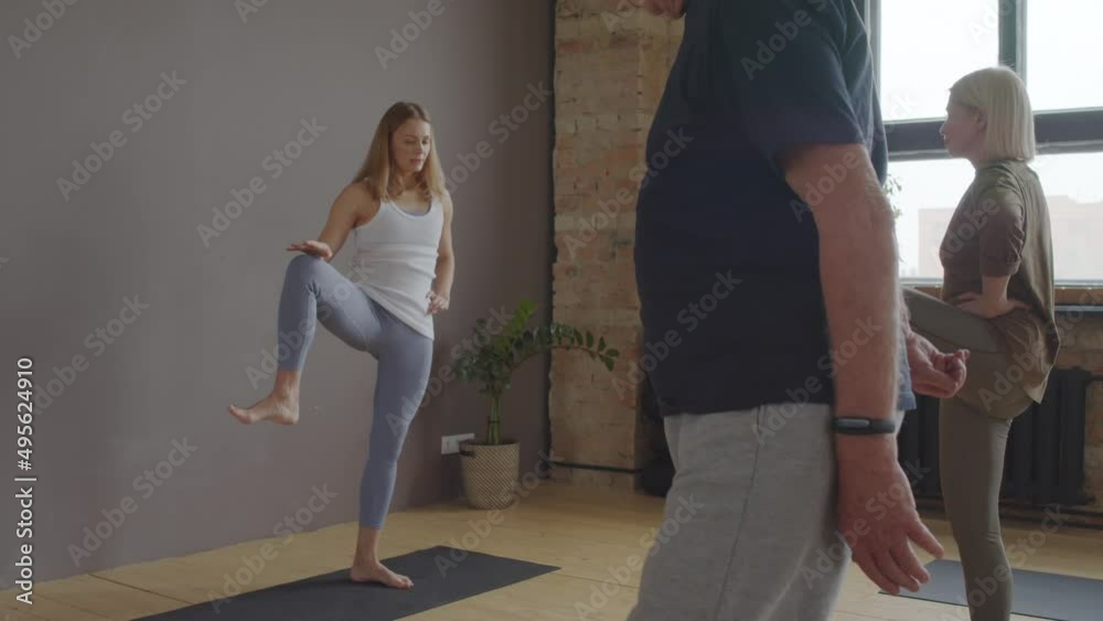 Young blond-haired female yoga instructor showing hip warming up exercise to senior man and woman in class at daytime Young blond-haired female yoga instructor showing hip warming up exercise to senior man and woman in class at daytime