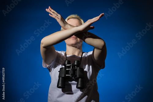 A blonde woman with binoculars around her neck shows a stop sign with her arms crossed on a dark background.