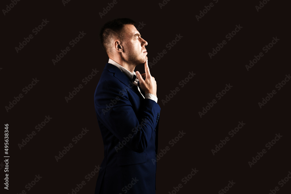Young male businessman in a blue suit stands sideways on a dark background with copy space in hard light