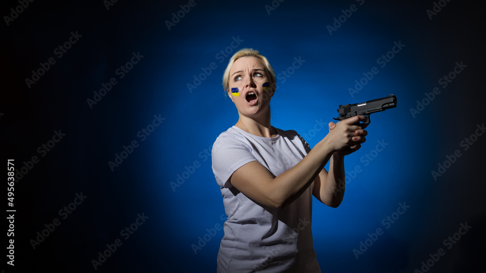 Aggressive woman in a white t-shirt screaming with a gun in her hands with the Ukrainian flag on her cheek on a dark background with copy space. Patriot concept. Aggressive woman in a white t-shirt screaming with a gun in her hands with the Ukrainian flag on her cheek on a dark background with copy space. Patriot concept.