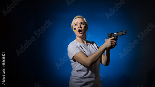 Aggressive woman in a white t-shirt screaming with a gun in her hands with the Ukrainian flag on her cheek on a dark background with copy space. Patriot concept.