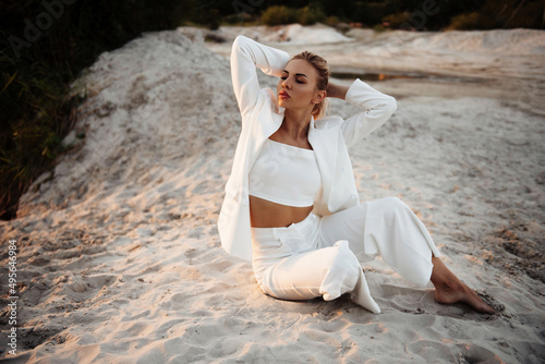 Fashionable lady in white suit sitting on sand and looking aside