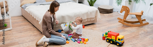 Woman looking at baby son holding bag with building blocks in bedroom, banner.