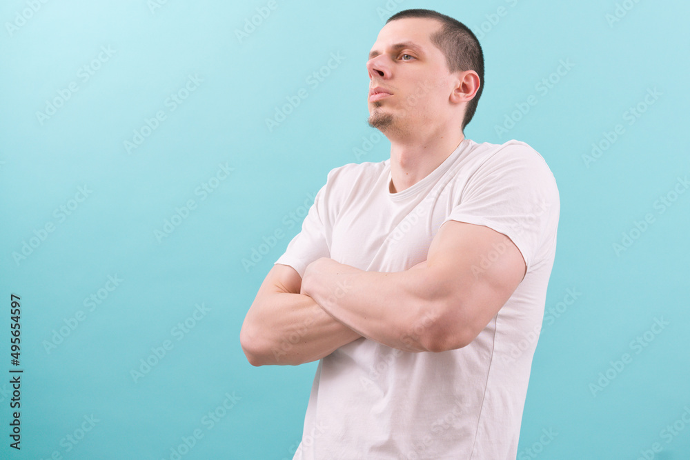 A caucasian strong man in a white t-shirt is standing confidently with crossed hands on a blue background. Person. Male. Man. Guy. Model. Studio. Young. Standing. Portrait. Attractive. Casual. Hand A caucasian strong man in a white t-shirt is standing confidently with crossed hands on a blue background. Person. Male. Man. Guy. Model. Studio. Young. Standing. Portrait. Attractive. Casual. Hand