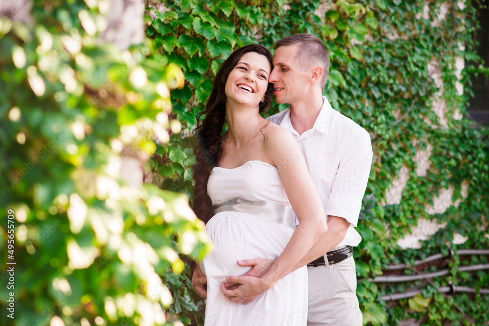 Young couple, Caucasian man and his pregnant wife in a field. Happy family resting on the wild field or meadow in summer. Man and pregnant woman in white dress and hat hugging belly Young couple, Caucasian man and his pregnant wife in a field. Happy family resting on the wild field or meadow in summer. Man and pregnant woman in white dress and hat hugging belly