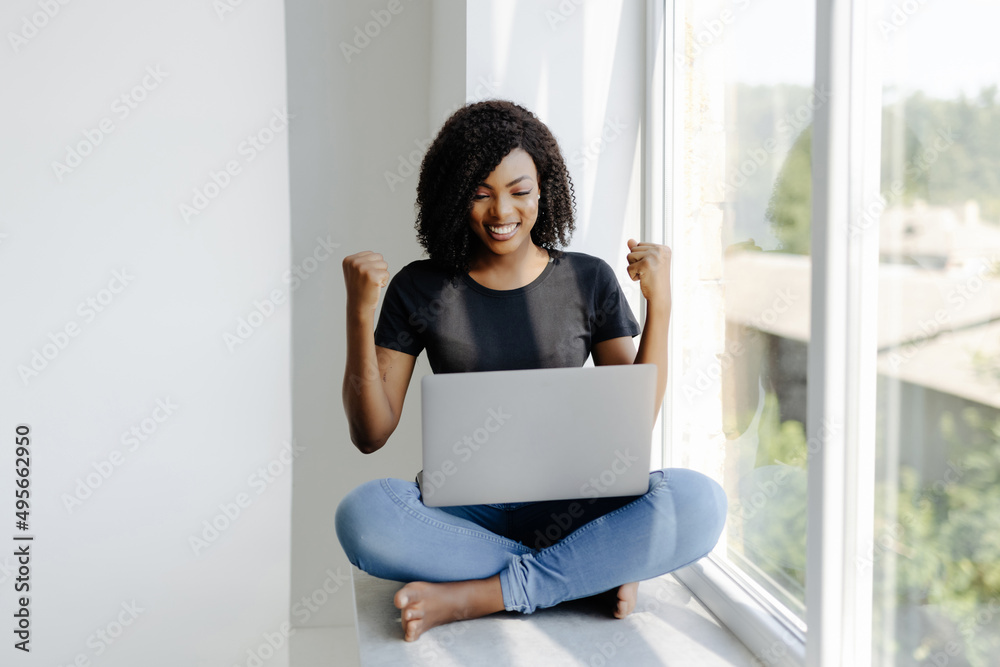 Relaxed African Americanwoman with laptop celebrate win sitting on windowsill Relaxed African Americanwoman with laptop celebrate win sitting on windowsill