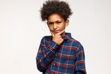 Picture of serious teen dark-skinned kid with cute stylish afro haircut holding chin with fingers, frowning, looking at camera with suspicious face expression, dressed in trendy flannel shirt
