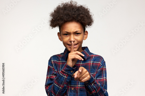 Frustrated dark-skinned handsome male kid in plaid shirt pointing at camera putting right index finger at mouth, asking for silence, hush, isolated against copy space for advertising content