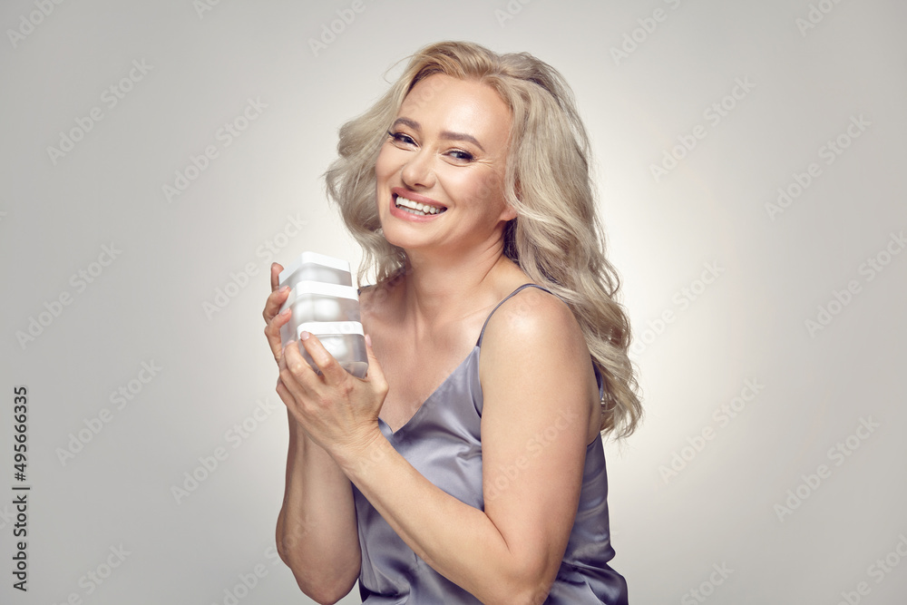 Senior blonde woman holding multiple jars of cream for face moisturising on grey isolated. Senior blonde woman holding multiple jars of cream for face moisturising on grey isolated.