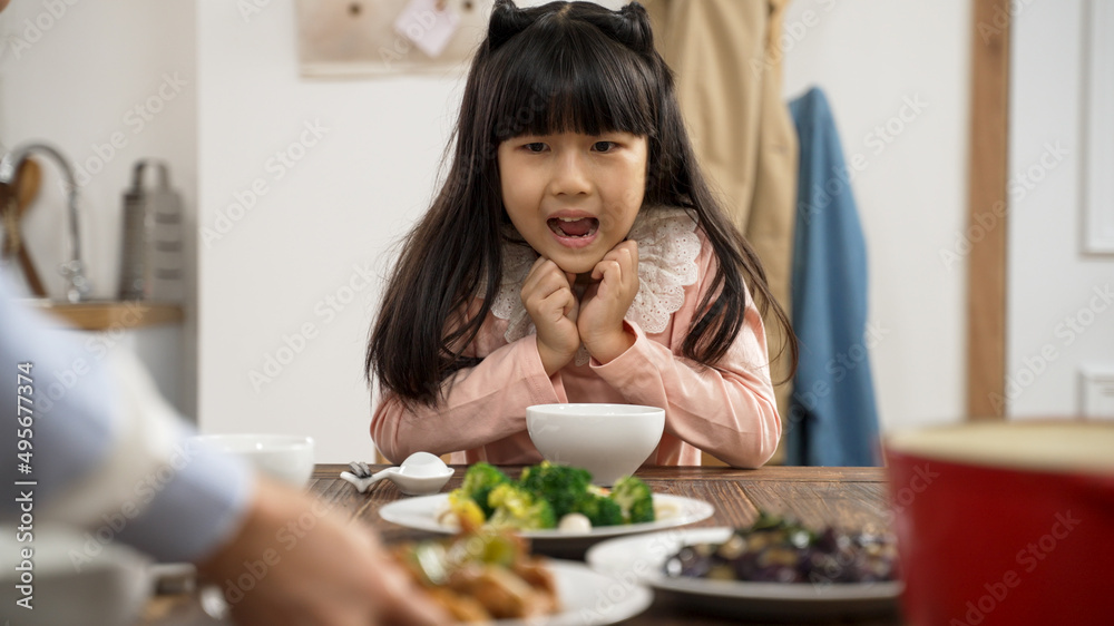selective focus of amazed Asian daughter holding face with anticipation her mother is putting on delicious dish on dinner table at home selective focus of amazed Asian daughter holding face with anticipation her mother is putting on delicious dish on dinner table at home