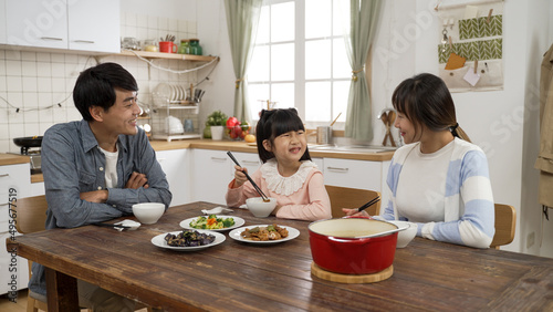 caring asian parents looking at their schoolgirl daughter eating at lunch table in a modern dining room with daylight. mom filling girl’s bowl with food and telling her to eat more